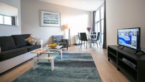 Een moderne woonkamer met een bank, salontafel en televisie. Er is een eethoek met een tafel en stoelen, en een groot raam zorgt voor natuurlijk licht.