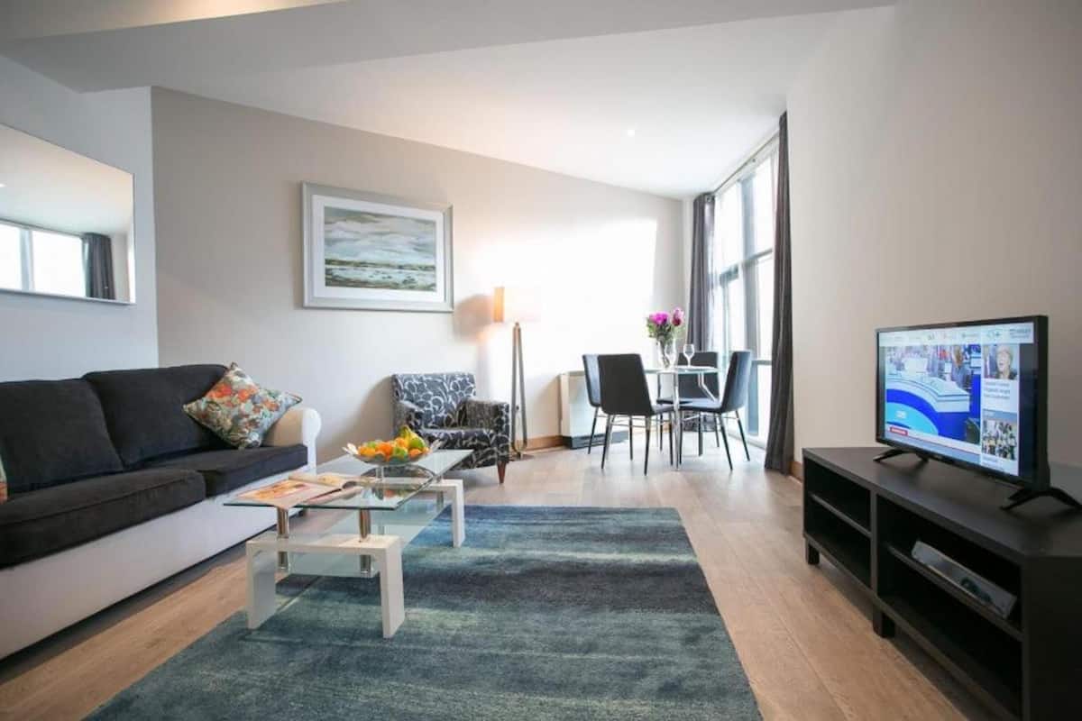 Una sala de estar moderna con sofá, mesa de centro y televisor. Hay un comedor con mesa y sillas, y un gran ventanal que proporciona luz natural.