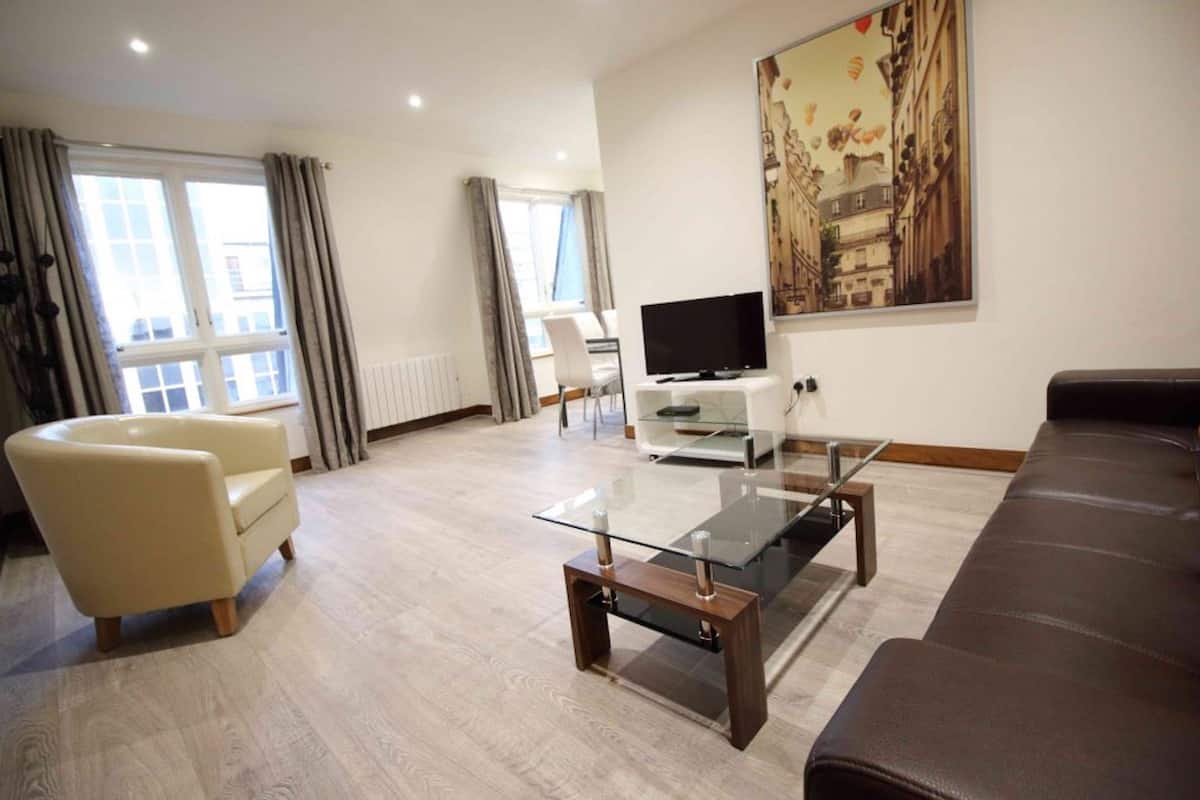 Una sala de estar moderna con una mesa de centro de vidrio, un sofá color beige, un sofá cama de cuero marrón y un televisor de pantalla plana.