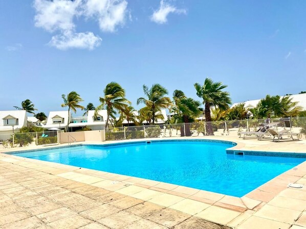 Una piscina con agua cristalina azul, rodeada de palmeras y un edificio blanco al fondo.