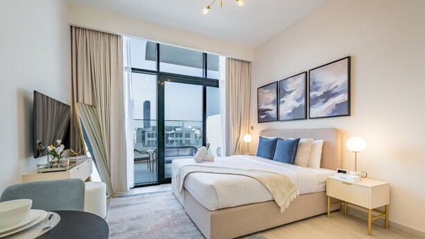 A modern hotel room with a large bed, a desk with a chair, a dining table with plates, and a view of the city through large windows.
