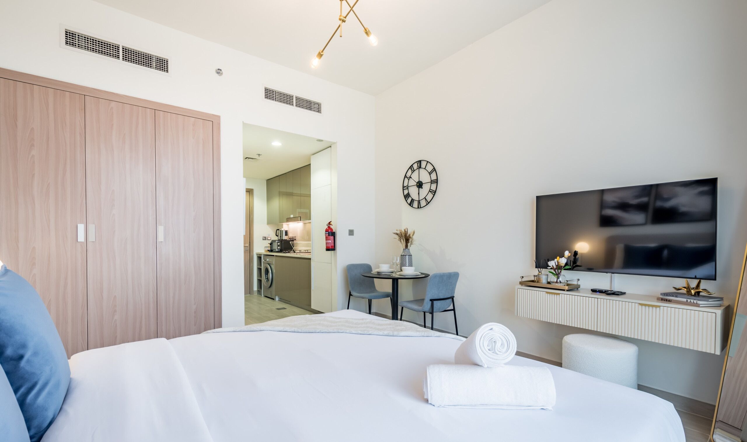 A modern hotel room with a bed, a desk with a flat-screen TV, a dining area, and a visible kitchen.