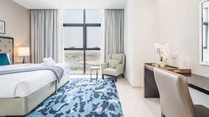 A hotel room with a bed, a desk, a chair, a small table, and a view of the cityscape through large windows. - Cosmopolitan 1BR Apartment in Celestia B, Dubai South by Deluxe Holiday Homes (Dubai)