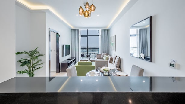 A modern hotel room with a dining area, a sofa set, a large window with a city view, and a chandelier. - Cosmopolitan 1BR Apartment in Celestia B, Dubai South by Deluxe Holiday Homes (Dubai)