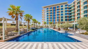 A swimming pool with lounge chairs and palm trees, surrounded by a modern building with balconies. - Cosmopolitan 1BR Apartment in Celestia B, Dubai South by Deluxe Holiday Homes (Dubai)