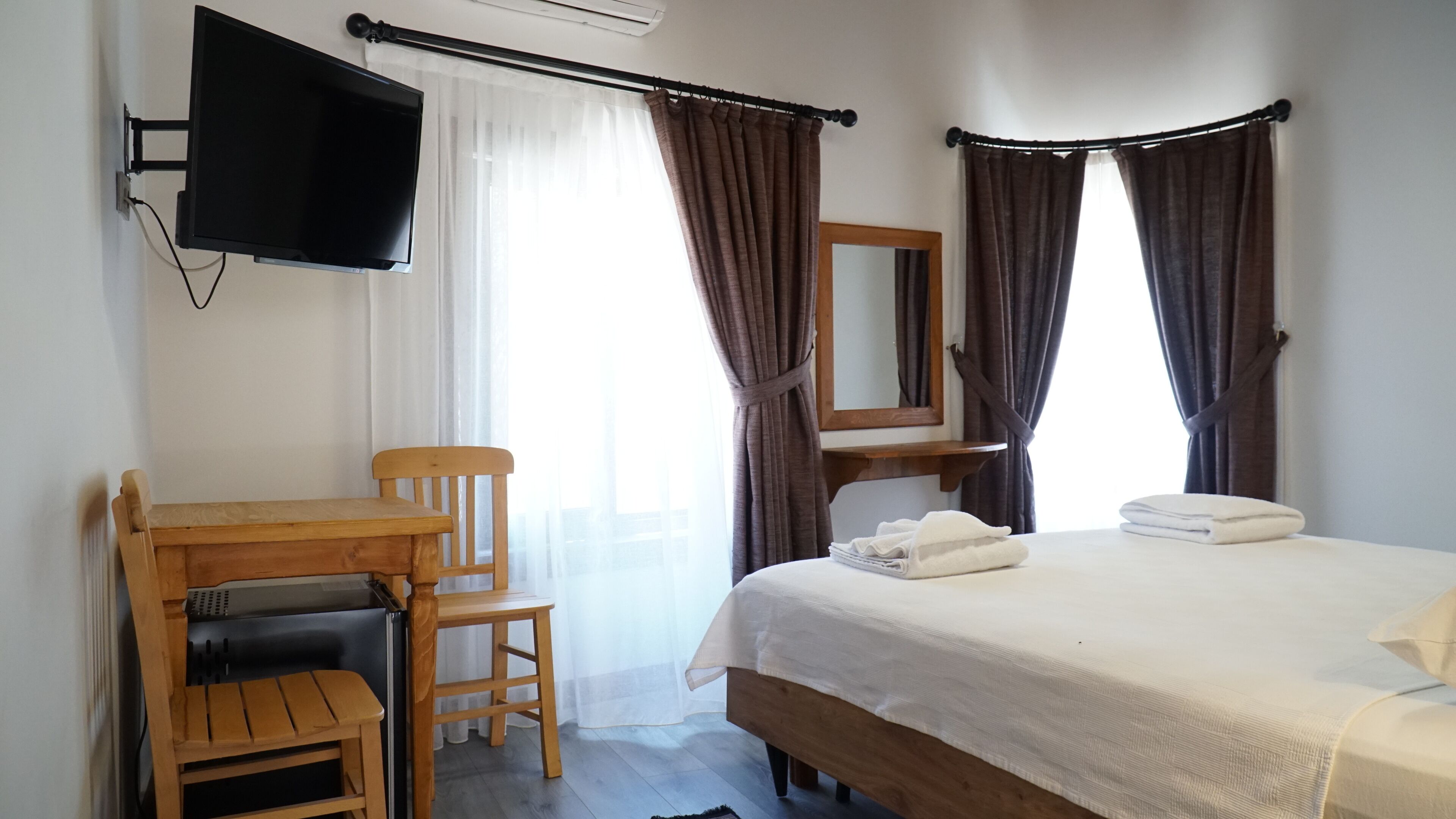 A hotel room with a bed, a desk with a chair, a television mounted on the wall, and a window with curtains.