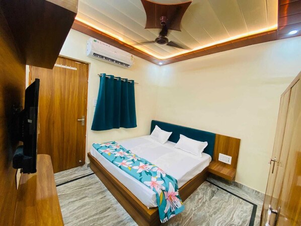 A bedroom with a bed, a ceiling fan, an air conditioning unit, a wooden wardrobe, and a television. - The Raman Palace - A Budget Hotel (Mathura)