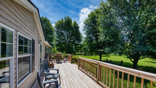A wooden deck with outdoor furniture, a view of a green landscape, and a house with large windows. - Trout House, Trout House 4bd/3ba on the South Holston River (Bristol)