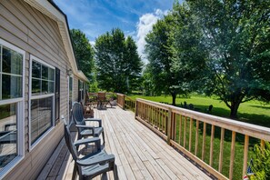 Une terrasse en bois avec des meubles d’extérieur, une vue sur un paysage verdoyant et une maison dotée de grandes fenêtres.