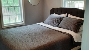 Ein Schlafzimmer mit einem Bett, zwei Fenstern mit Jalousien, einem runden Spiegel und einer Deckenleuchte.