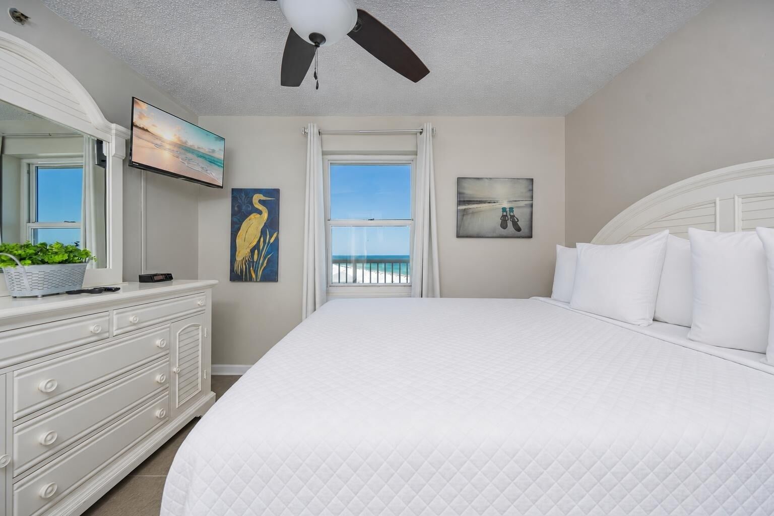 Une chambre avec un grand lit, un ventilateur de plafond, une commode, un téléviseur et une vue sur la plage.