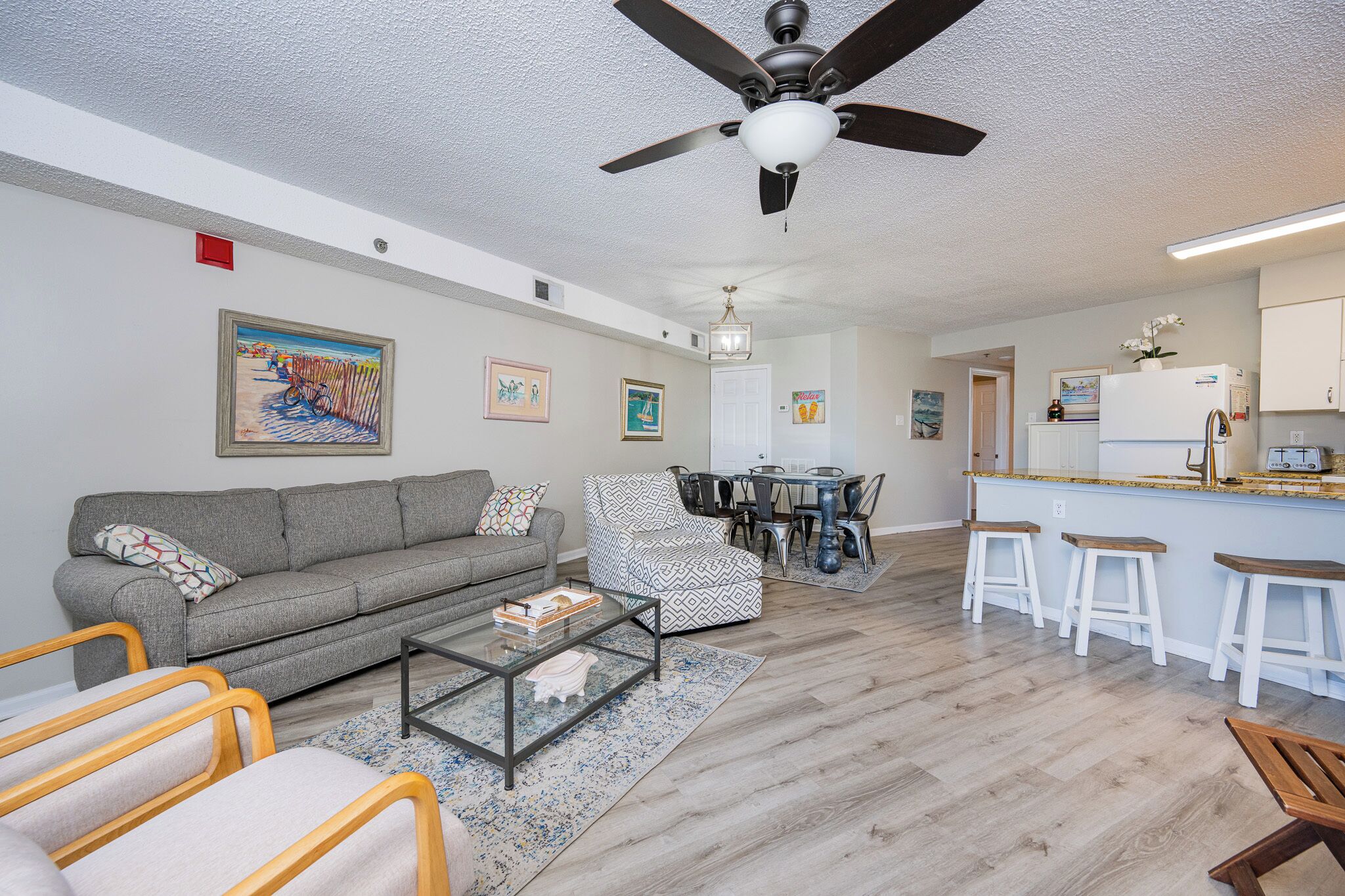 A living room with a sofa, coffee table, and patterned armchairs, a dining area with a table and chairs, and a kitchen with a refrigerator and bar stools.