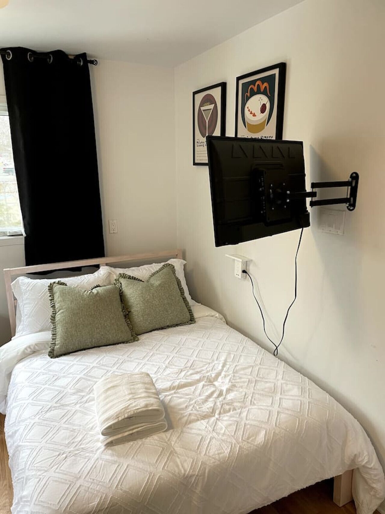 A neatly made bed with white bedding and two pillows, a black curtain on the window, and two framed pictures on the wall.