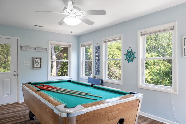 A pool table in a room with large windows, a ceiling fan, and a nautical-themed wall decoration.