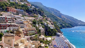 Une ville côtière avec des bâtiments construits sur une colline, une plage parsemée de parasols et des bateaux sur l’eau.