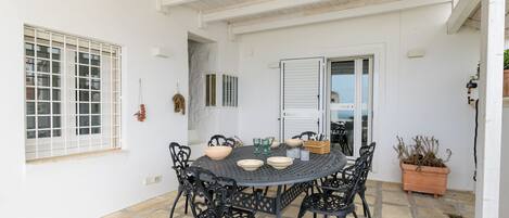 Un porche cubierto con una mesa redonda y sillas, una planta en maceta y una ventana con contraventanas.