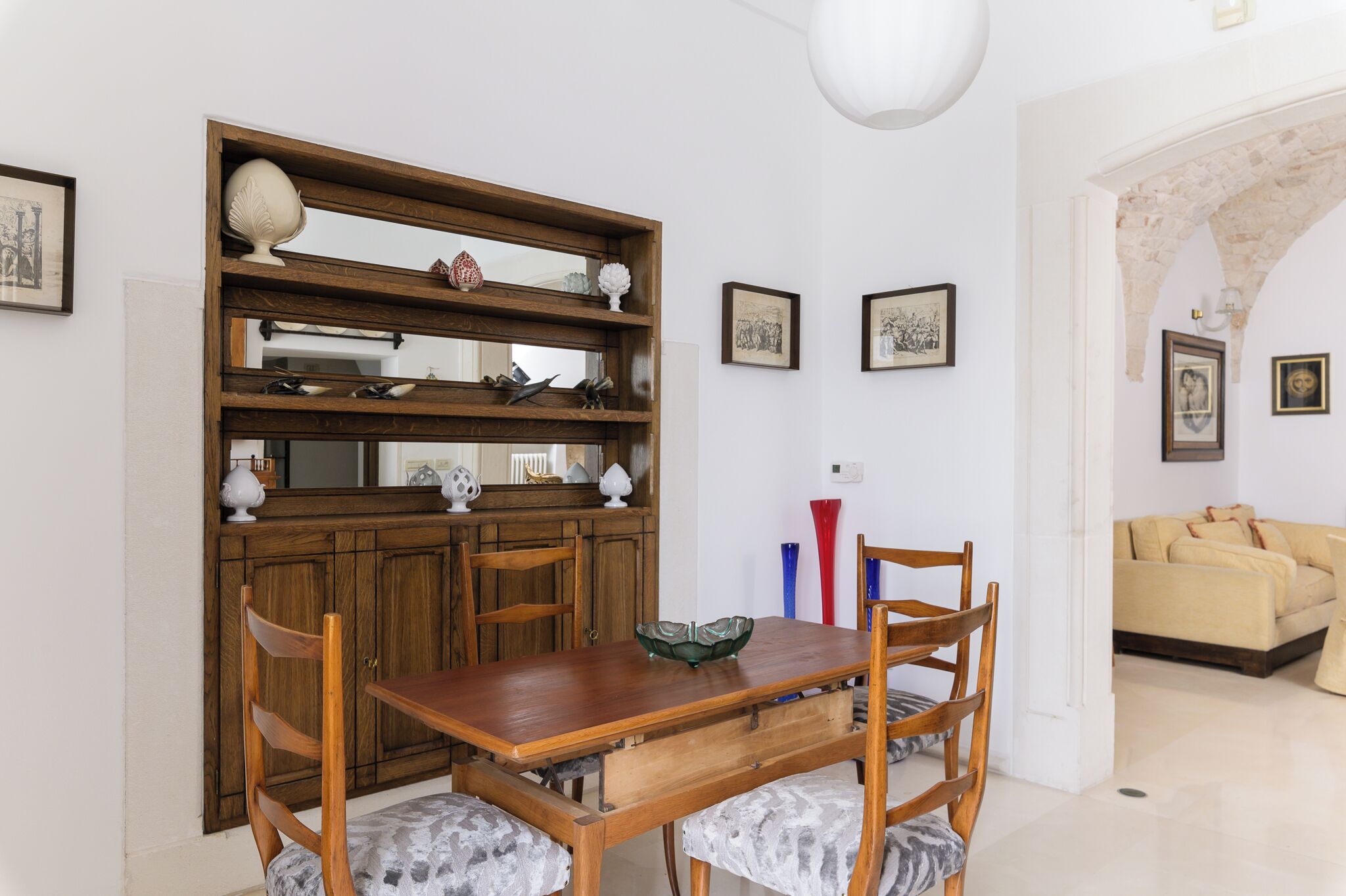 Un comedor con una mesa y sillas de madera, un estante decorativo con diversos objetos, y un sofá al fondo.