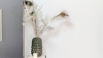 En dekorativ vase med påfugle fjer og tørrede planter på en hvid hylde foran en grå væg.