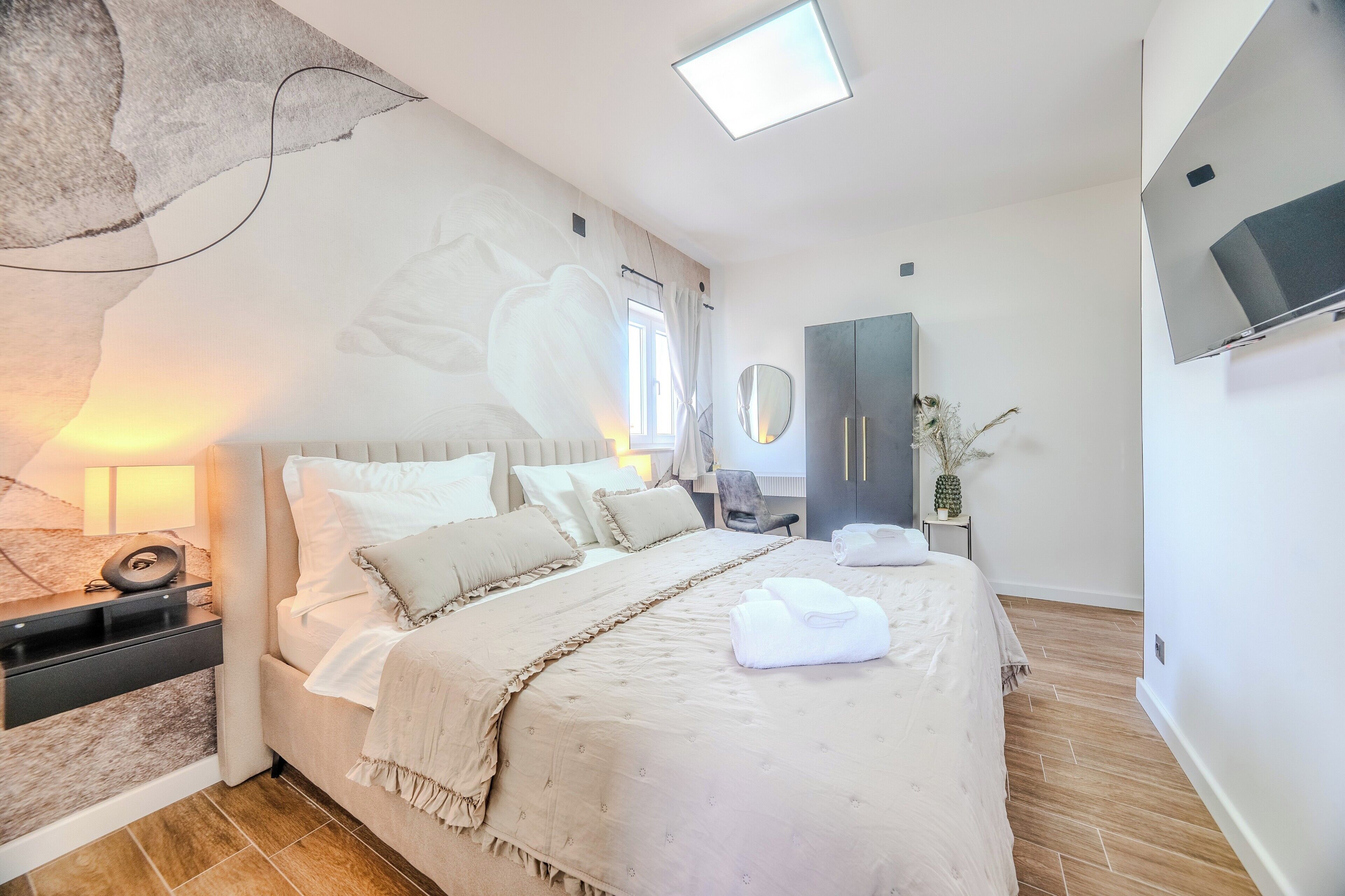 A modern bedroom with a bed, bedside table, mirror, wardrobe, and a flat-screen TV.