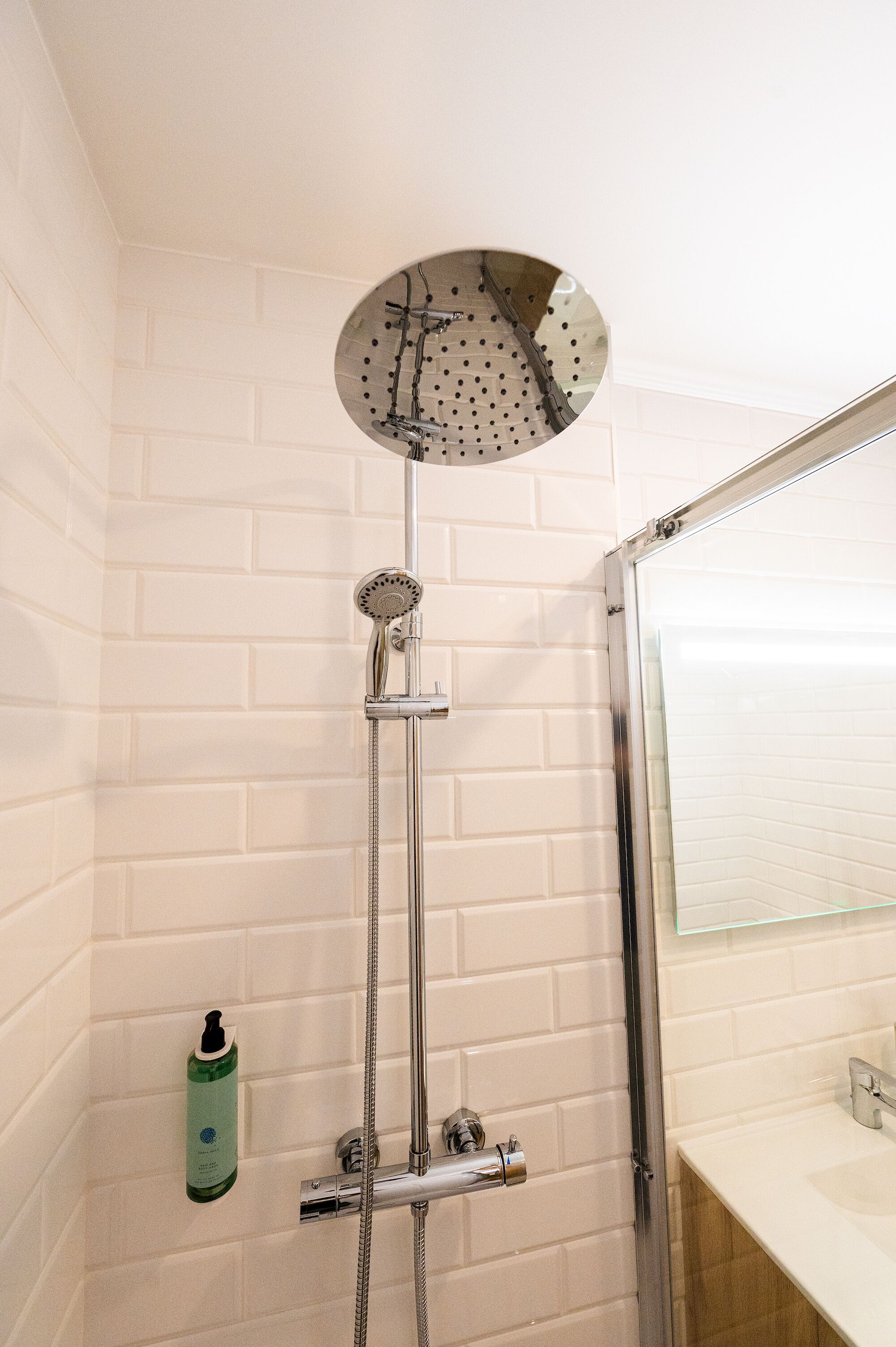 A shower with a handheld showerhead and a wall-mounted faucet, a bottle of shampoo, and a mirror above the sink.