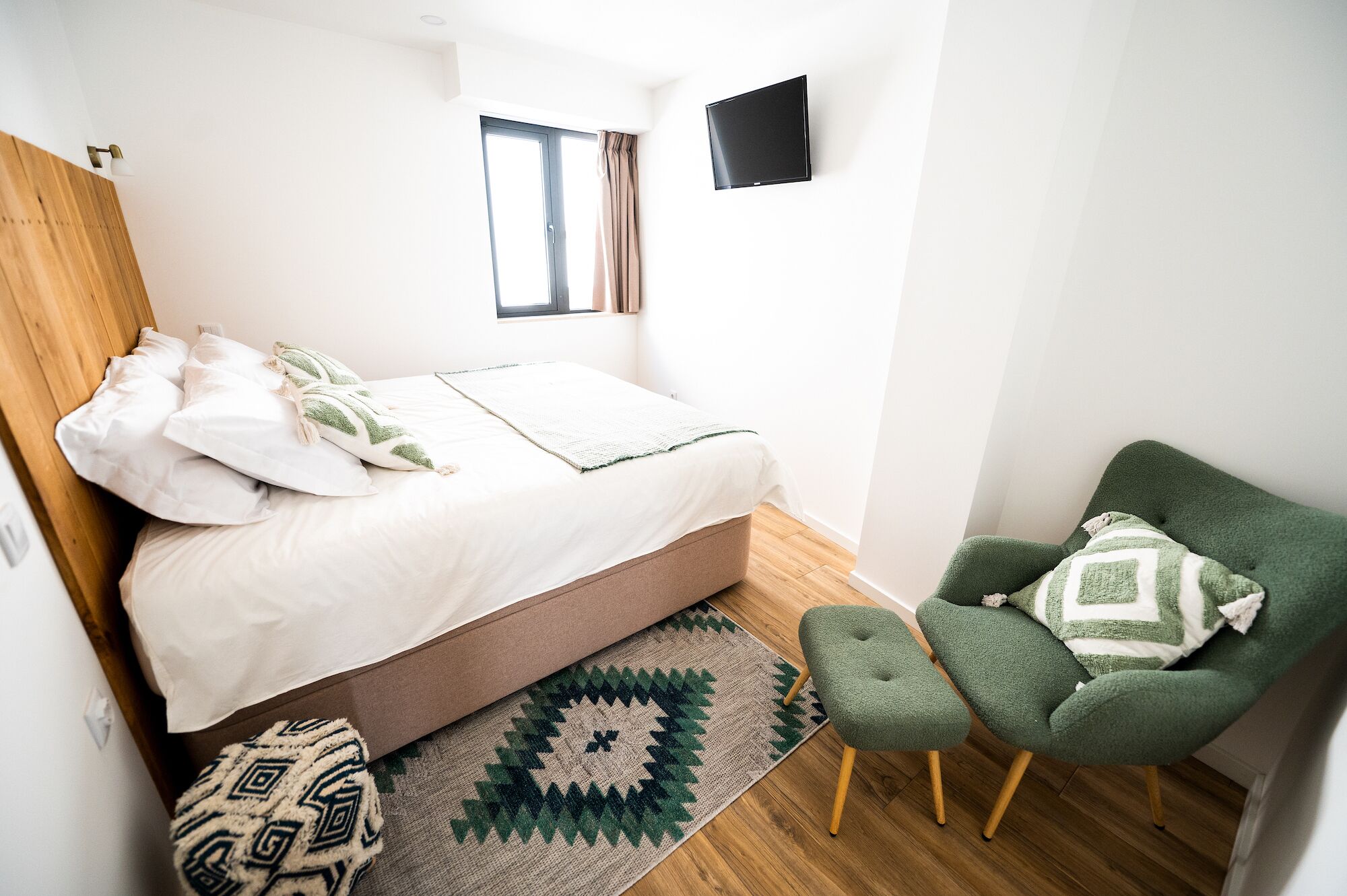 A bedroom with a bed, a green armchair, a TV, and a window.