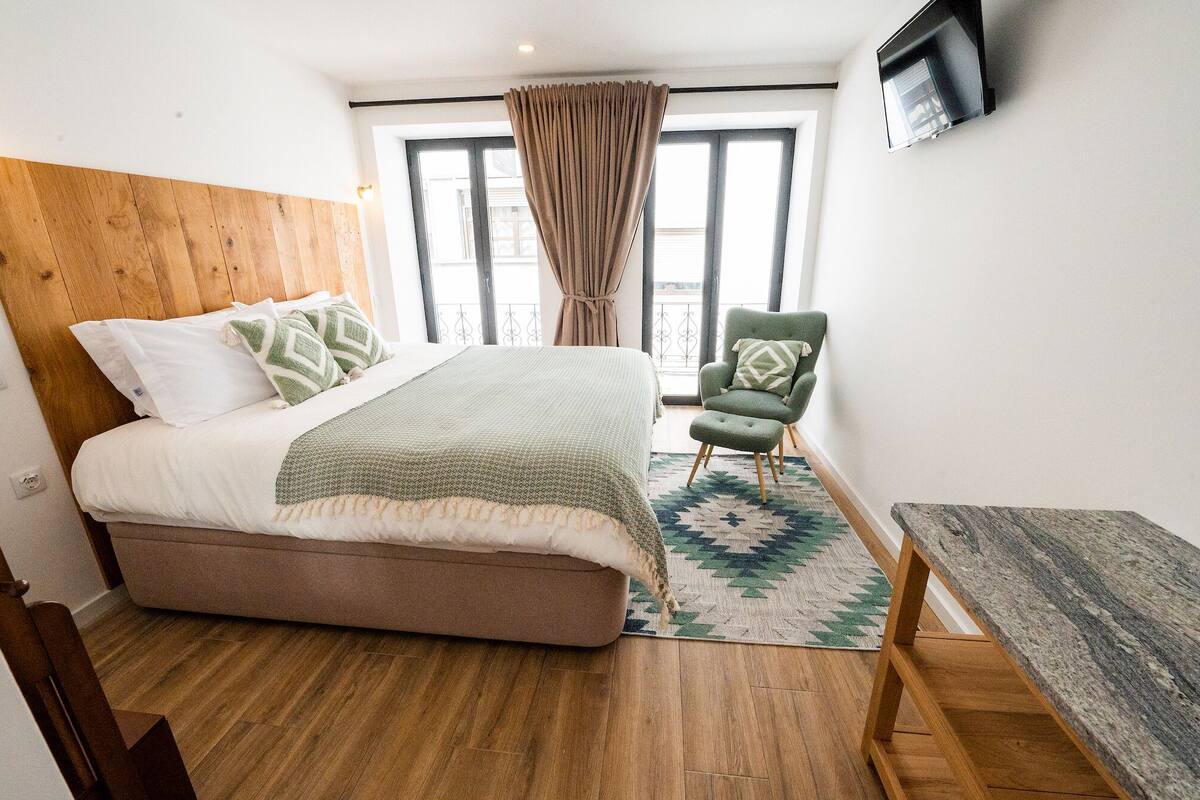 A bedroom with a wooden headboard, a bed with patterned bedding, a green armchair, a small table, and a TV mounted on the wall.