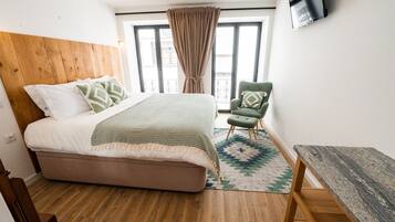 A bedroom with a wooden headboard, a bed with patterned bedding, a green armchair, a small table, and a TV mounted on the wall.