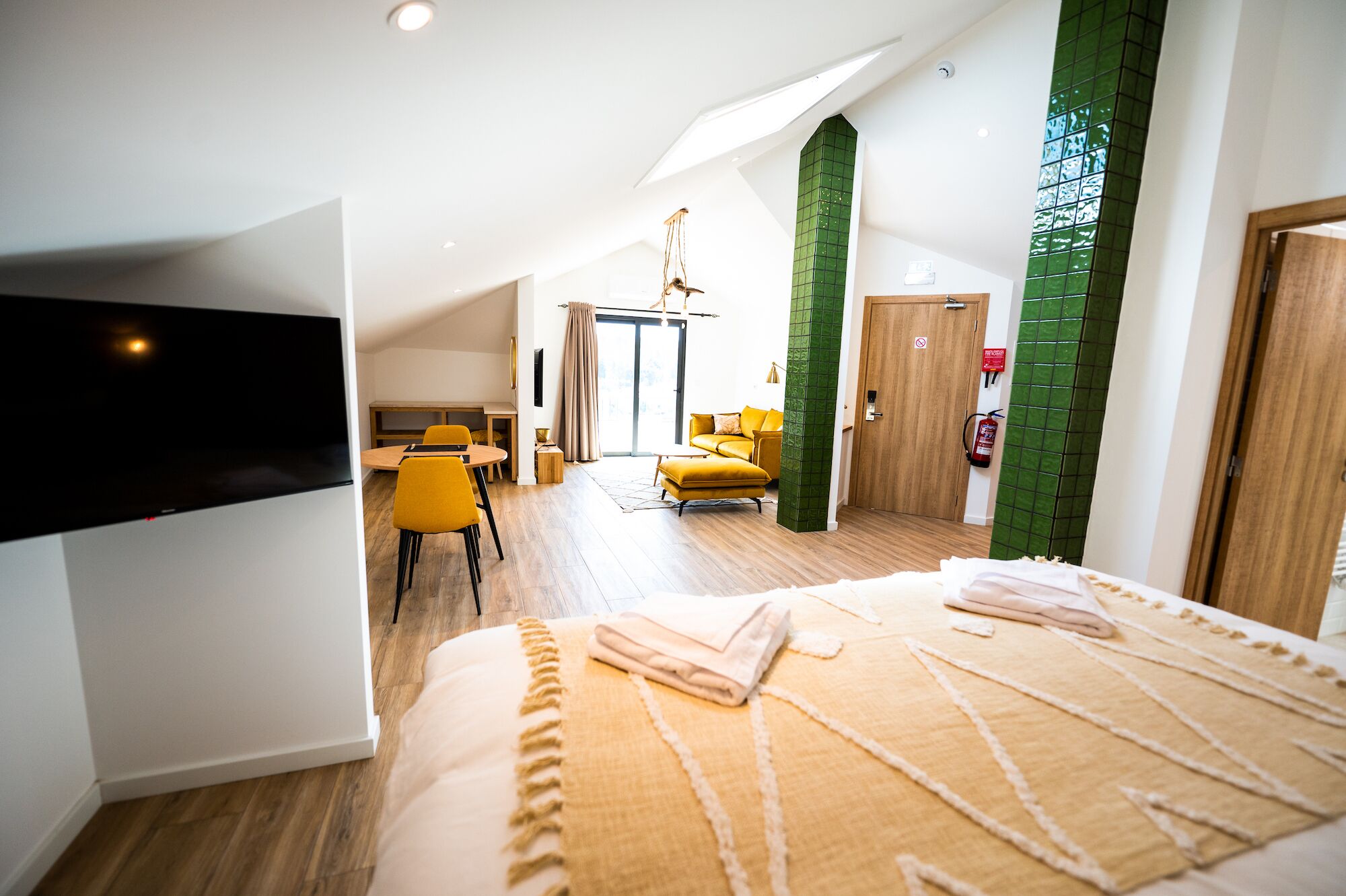 A modern hotel room with a large bed, a dining area with yellow chairs, and a green accent wall.