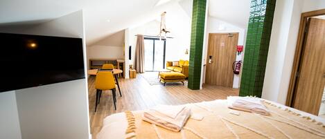 A modern hotel room with a large bed, a dining area with yellow chairs, and a green accent wall.