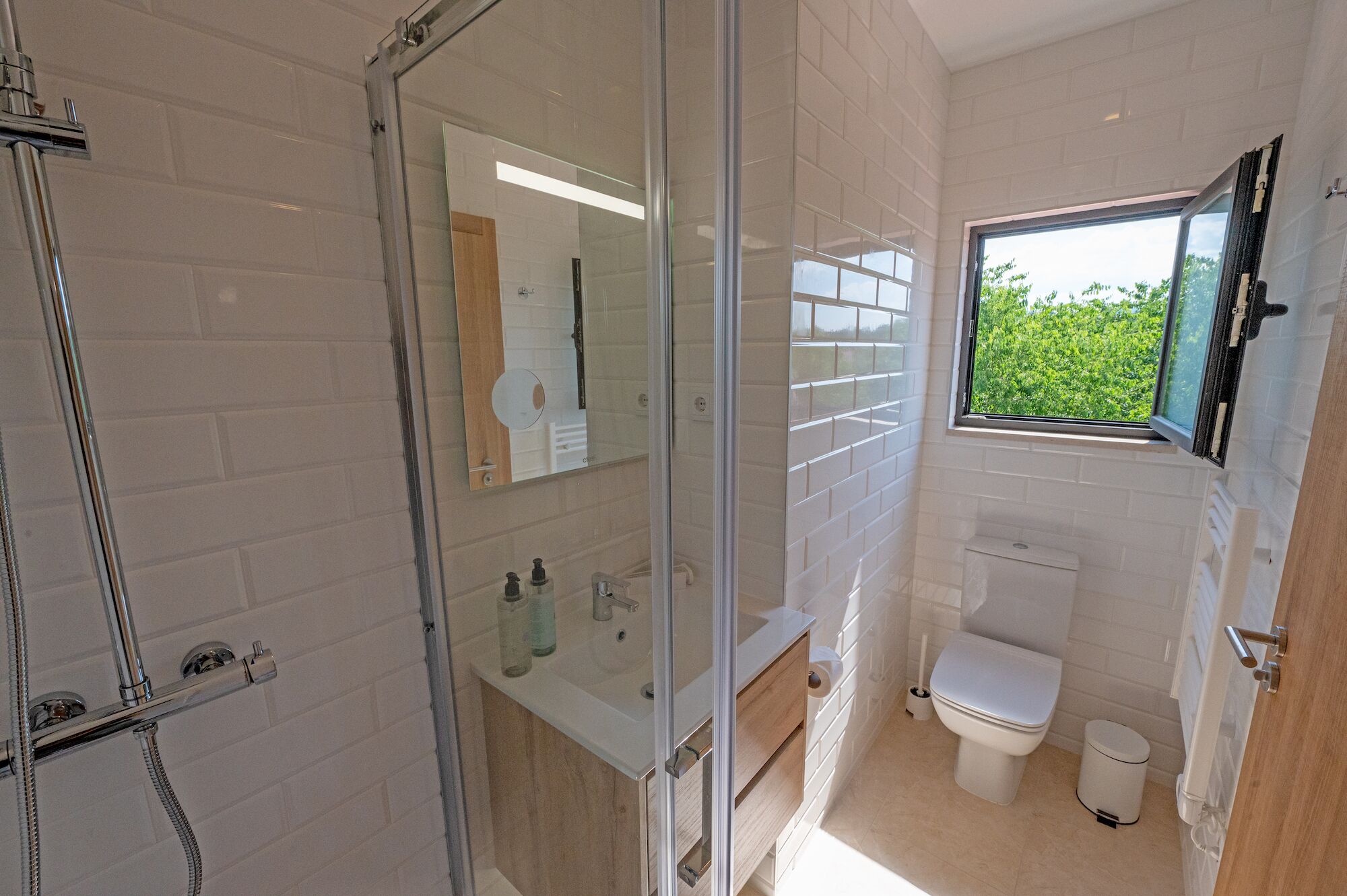 Een moderne badkamer met een glazen douchecabine, een toilet en een raam met uitzicht op groen.