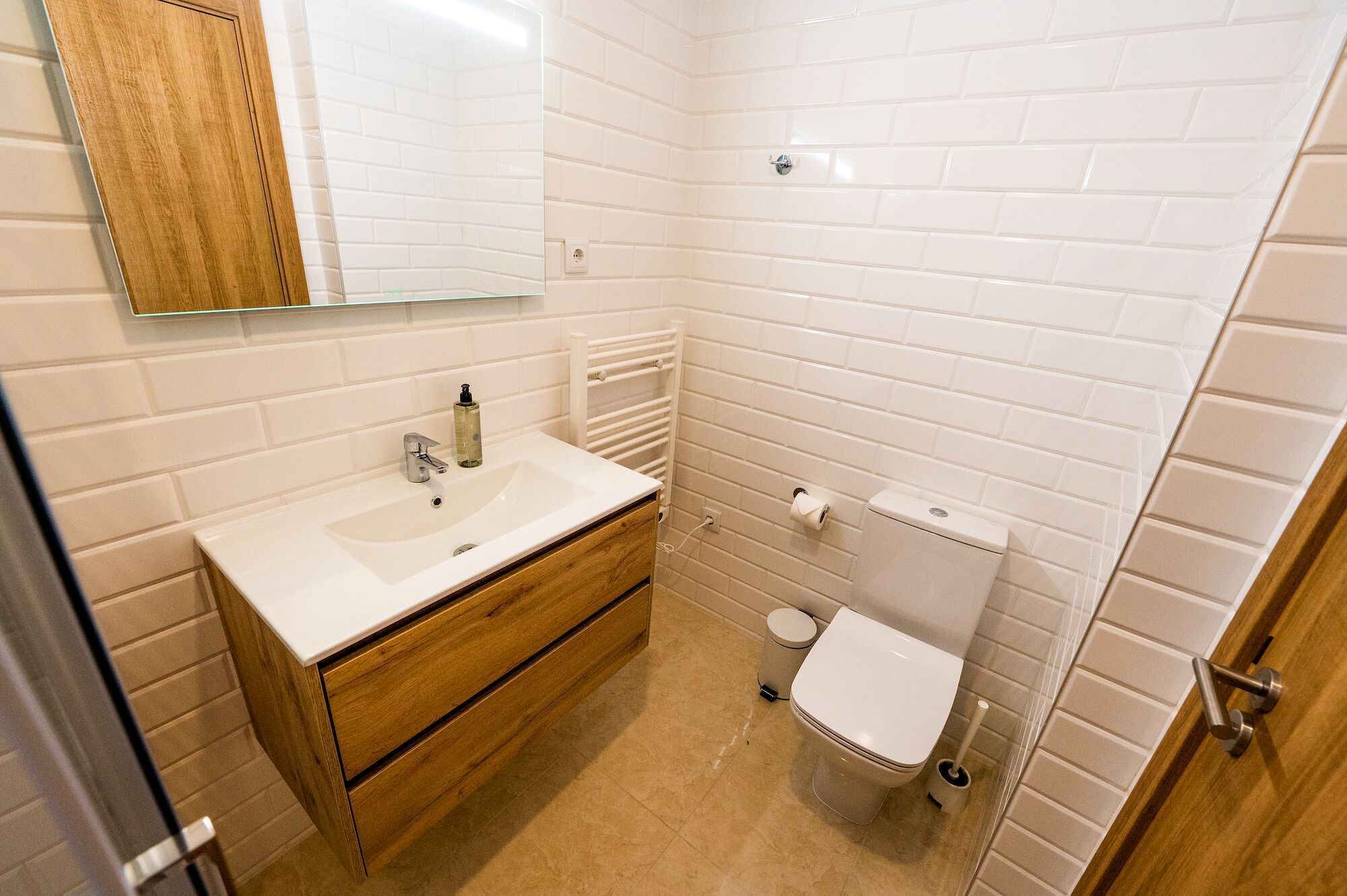 Un baño pequeño y moderno con azulejos blancos tipo metro, un mueble de tocador de madera con lavabo, un espejo y un inodoro.