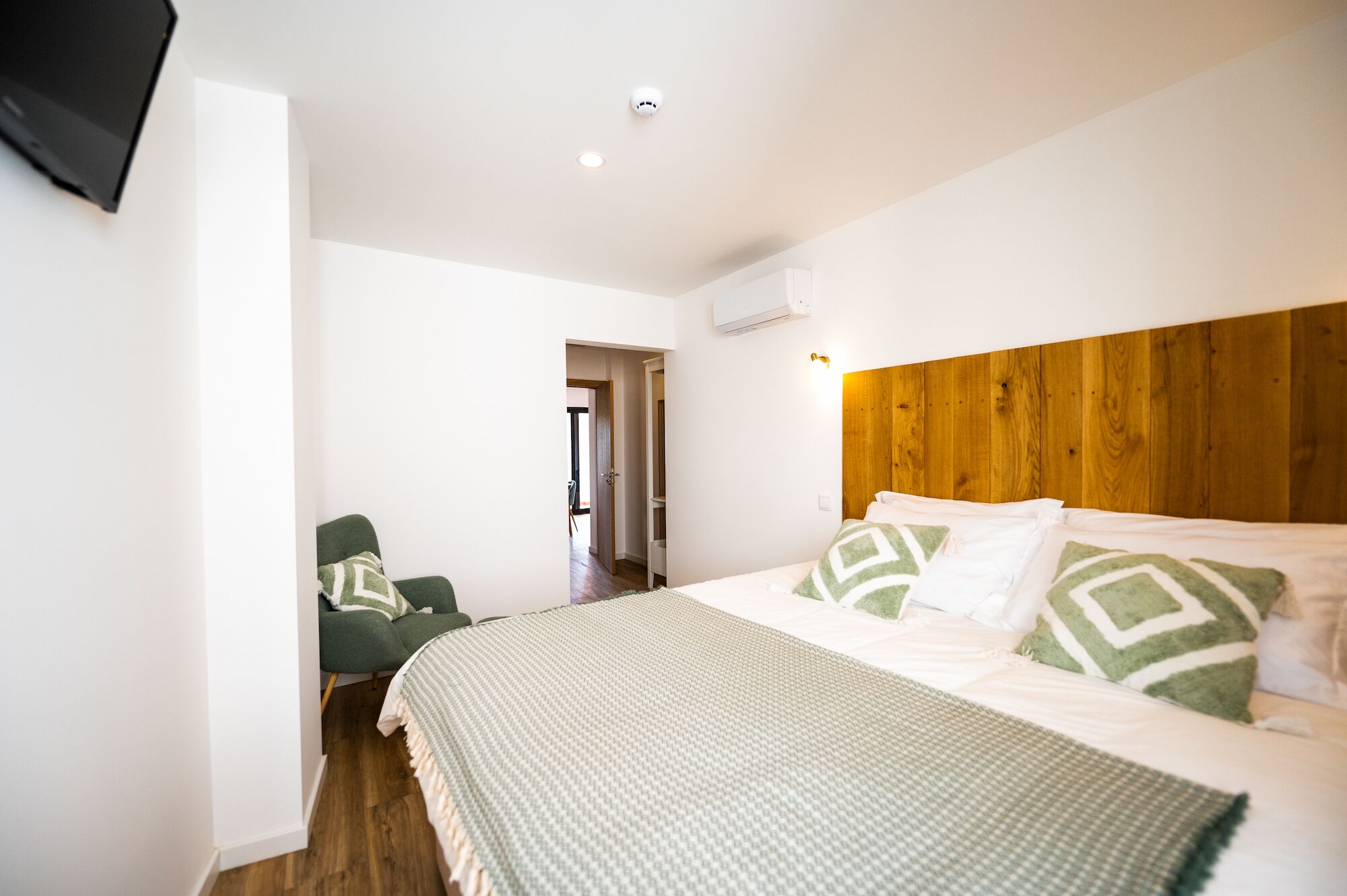 A bedroom with a wooden headboard, a bed with patterned bedding, a green armchair, and a television mounted on the wall.