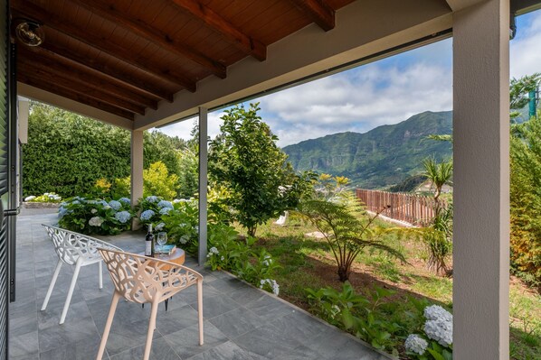 An outdoor patio with a view of mountains, a wooden pergola, and a garden with white flowers and plants.