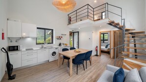 Une cuisine moderne avec des meubles blancs, un coin repas avec une table en bois et des chaises bleues, un escalier menant à une mezzanine, et une vue sur la verdure à l’extérieur.