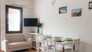A dining area with a table set for two, a sofa, a TV, and a window with blinds.