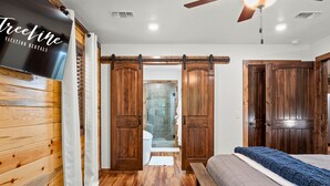 A room with wooden walls, a bed, a ceiling fan, and a sliding door leading to a bathroom. - Pet Friendly Woodland Escape with Play Area (Broken Bow)