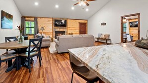 A spacious living area with a wooden wall, a fireplace, a sofa, and a dining table. - Pet Friendly Woodland Escape with Play Area (Broken Bow)