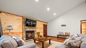 A cozy living room with a fireplace, a wooden accent wall, and a sign for 'Treeline Vacation Rentals'. - Pet Friendly Woodland Escape with Play Area (Broken Bow)