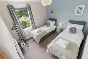 A bedroom with two single beds, a window with curtains, a bedside table with a lamp, and a 'HOME' sign on the wall.