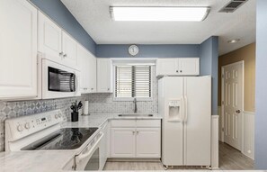 A kitchen with white cabinets, a microwave, a stove, a refrigerator, and a window with blinds.