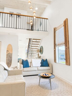 A spacious living area with a high ceiling, a staircase, a sofa set, a coffee table, and a surfboard decoration.