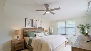 Un dormitorio con cama, mesitas de noche, ventilador de techo, ventana con persianas y una planta.