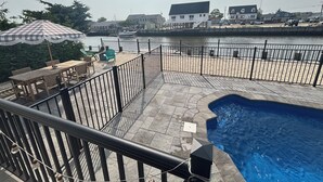 Un espace piscine avec vue sur une rivière, une terrasse en bois avec des chaises et un parasol à carreaux.