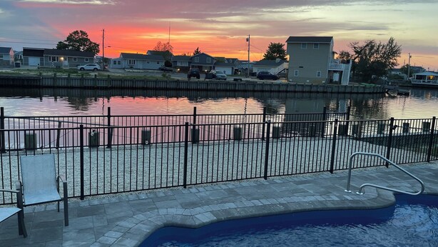 Une piscine entourée d’une clôture en métal et dotée d’une jetée s’avançant dans l’eau, le tout bordé de bâtiments résidentiels et surplombé par un ciel au coucher du soleil.