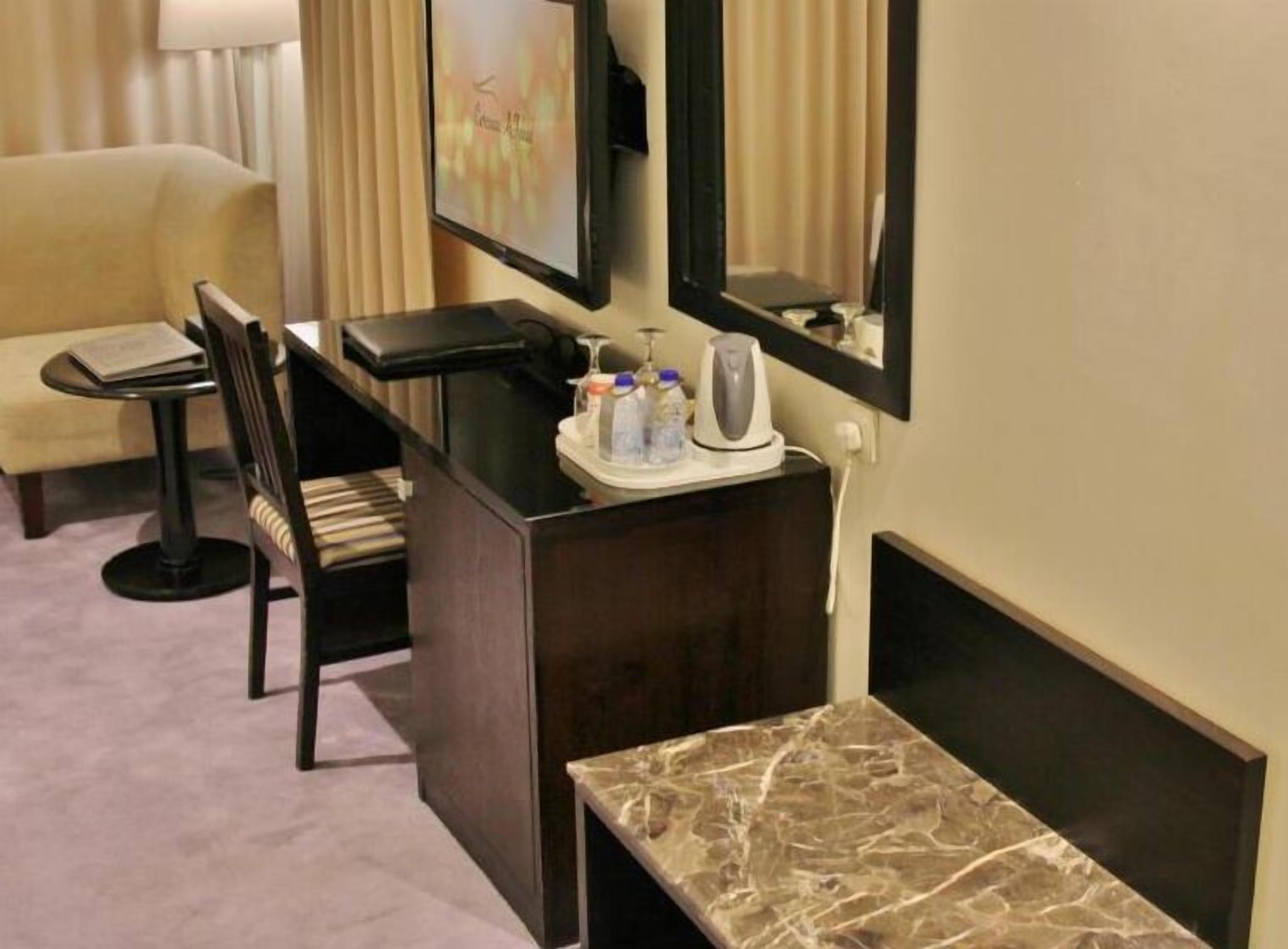 A hotel room with a desk, chair, and a small table. There is a mirror above the desk and a television mounted on the wall.