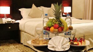 Ein Hotelzimmer mit einem ordentlich bezogenen Bett, einer Obstschale auf dem Nachttisch und einem Tablett mit Gebäck und einem Servietttuch.