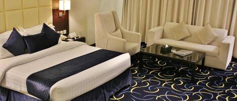 A hotel room with a bed, two armchairs, a sofa, a glass coffee table, and a patterned carpet.