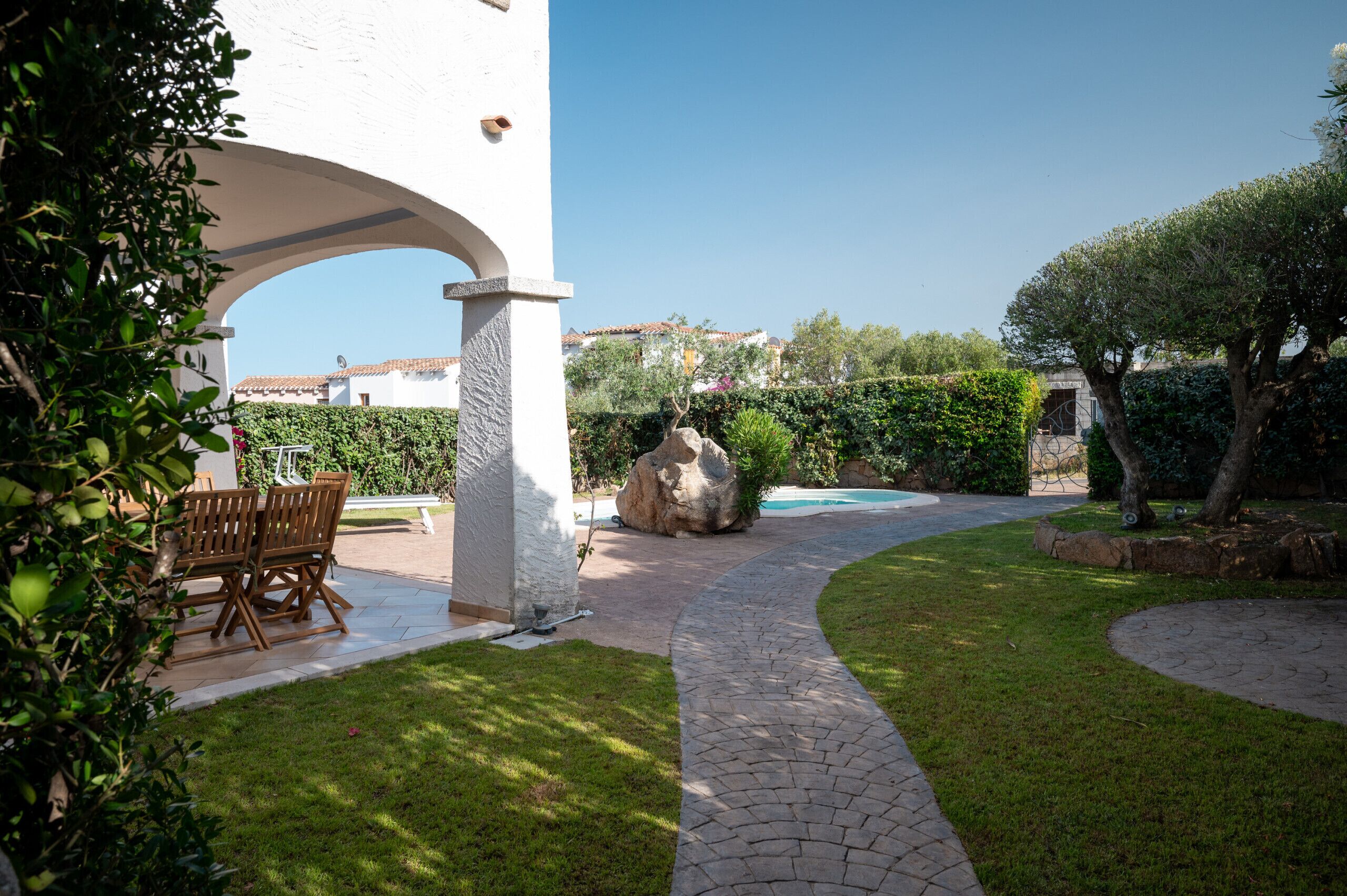 A patio area with a stone pathway, a wooden table and chairs, and a view of a swimming pool and buildings in the background.