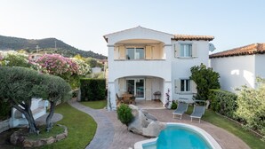 Una casa de dos pisos con piscina, un patio con mobiliario exterior y un jardín con diversas plantas y flores.