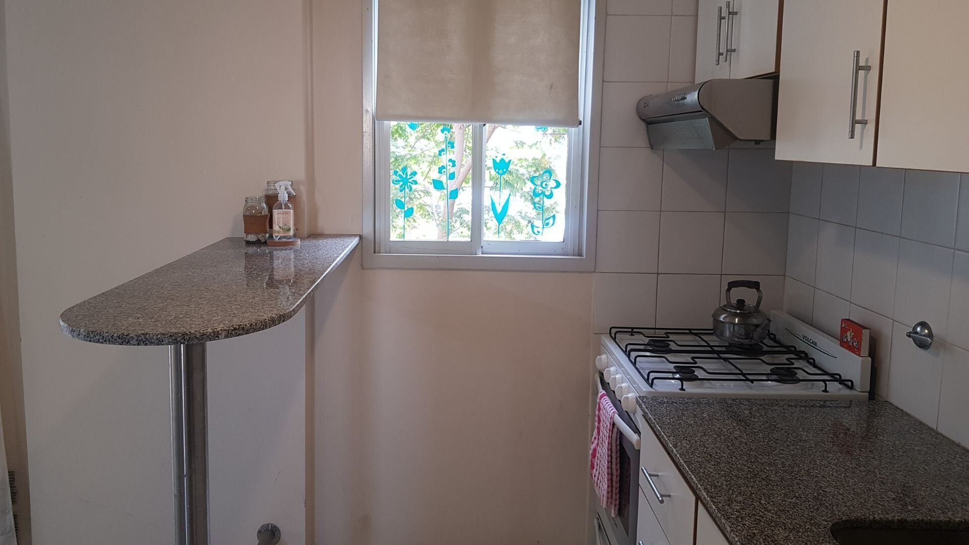 Een keuken met een gasfornuis, een raam met decoratieve blauwe bloemen en een kleine tafel met een fles en een pot.